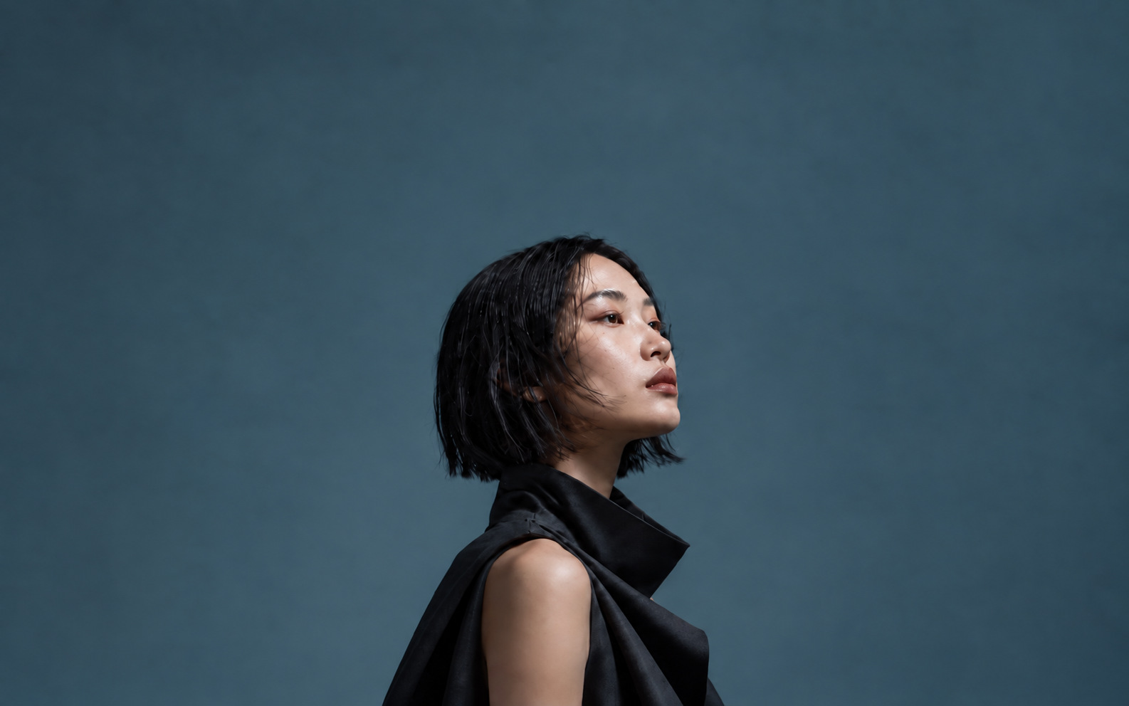 Portrait of a woman with a black bob in a sculptural black collar, looking up against a slate-blue wall
